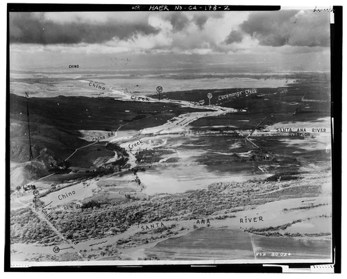  'SANTA ANA RIVER AT CHINO CREEK, RIVERSIDE COUNTY.' This is an oblique aerial view to the north, looking over the flooded fields between Chino Creek and the Santa Ana River, just upstream of the Prado Dam site. File number written on negative: R & H 80 024. - Prado Dam, Santa Ana River near junction of State Highways 71 & 91, Corona, Riverside County, CA