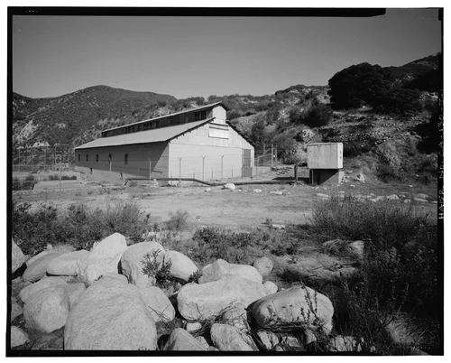  SAR-1, SOUTHEAST AND SOUTHWEST ELEVATIONS, WITH SWITCH RACK AT LEFT, AND SANTA ANA WELL #1 AND STONE RETAINING WALLS AT RIGHT. VIEW TO NORTH. - Santa Ana River Hydroelectric System, SAR-1 Powerhouse, Redlands, San Bernardino County, CA